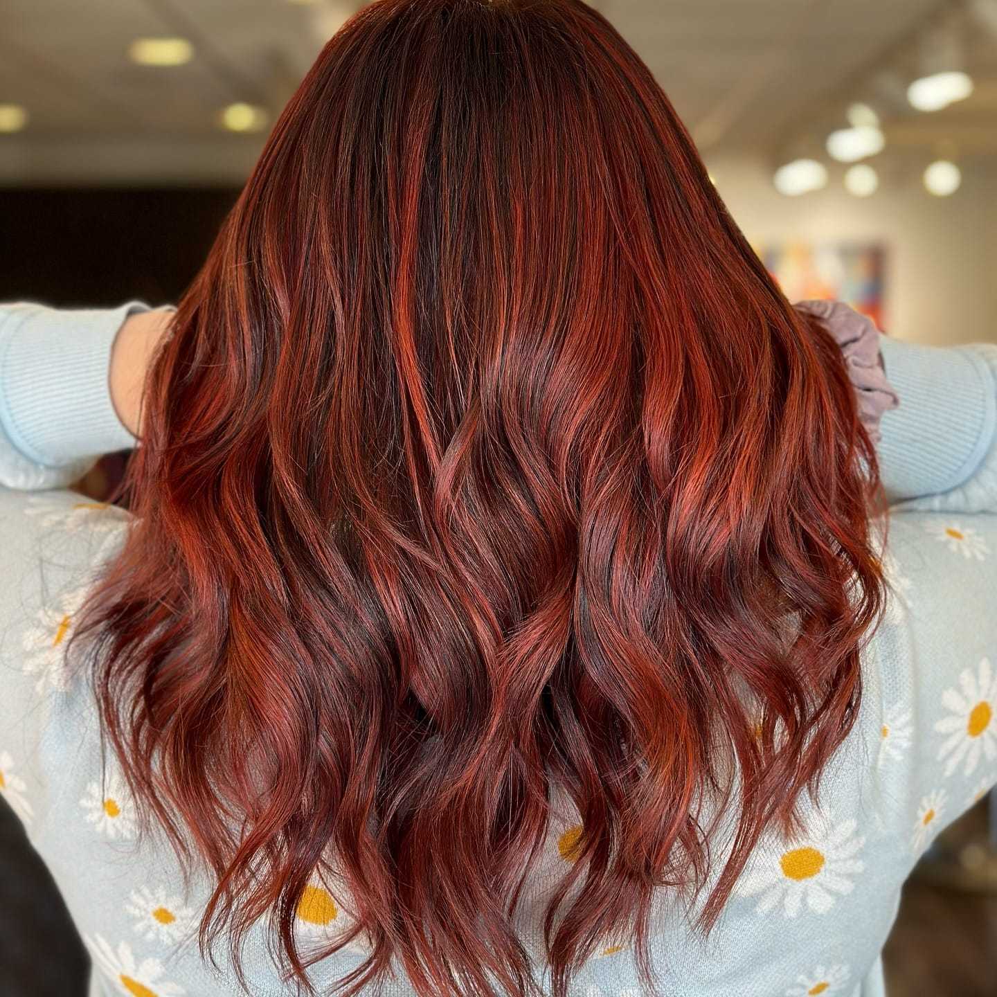 Wavy auburn hair cascading down the back, with a daisy-patterned sweater in soft lighting.
